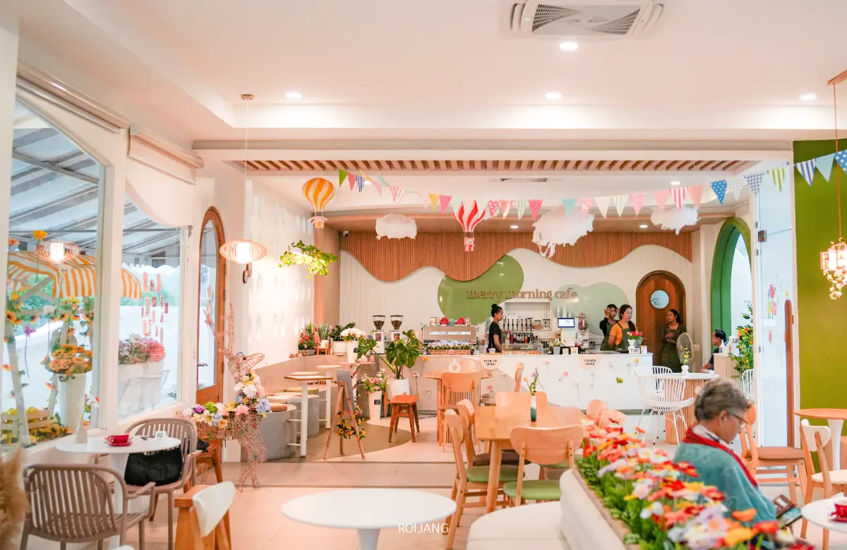 ภายในร้าน Merry Morning Cafe ที่สดใสและมีสีสัน มีทั้งของประดับแขวน ต้นไม้ และคนที่มาสั่งอาหารตรงเคาน์เตอร์ มีโต๊ะและเก้าอี้จัดวางอยู่ทั่วทั้งร้าน.