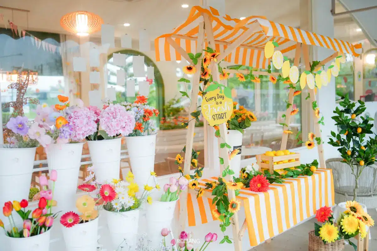 ร้านขายดอกไม้ที่มีหลังคาทรงแถบสีเหลืองและสีขาวจัดแสดงอยู่ภายในอาคารที่ Merry Morning Cafe รายล้อมไปด้วยกระถางสีขาวที่เต็มไปด้วยดอกไม้ประดิษฐ์หลากสีสันและของประดับดอกไม้.