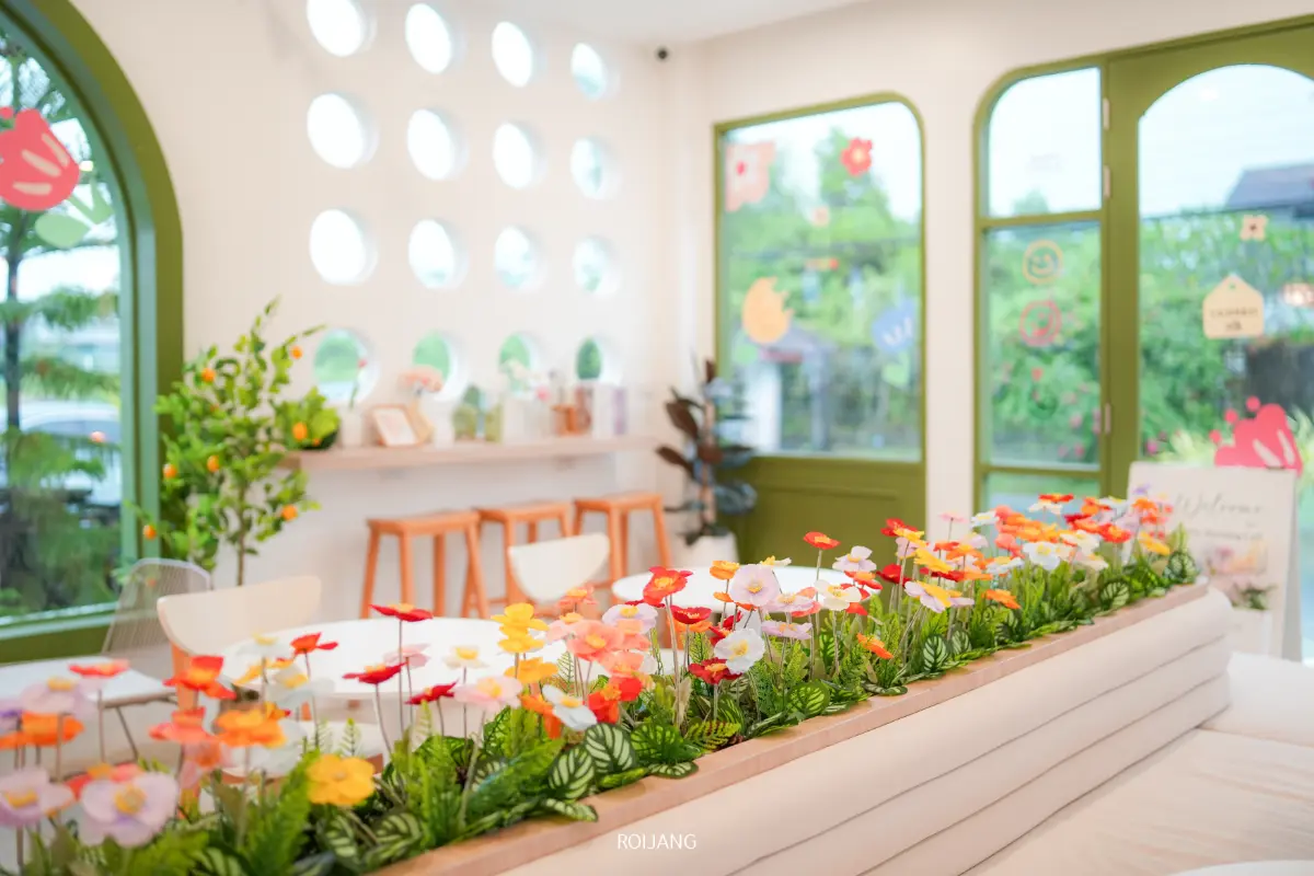 คาเฟ่ Merry Morning Cafe มีบรรยากาศภายในที่สดใสและทันสมัย โดดเด่นด้วยหน้าต่างทรงกลม ประตูกรอบสีเขียว พื้นที่นั่งที่บาร์ และดอกไม้ประดิษฐ์สีสันสดใสในกระถาง.