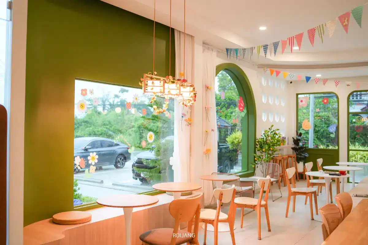 Merry Morning Cafe เป็นคาเฟ่ที่มีแสงสว่างสดใส มีผนังสีเขียว โต๊ะและเก้าอี้ไม้ หน้าต่างบานใหญ่ ไฟแขวน และธงรูปธงสีสันสดใส.