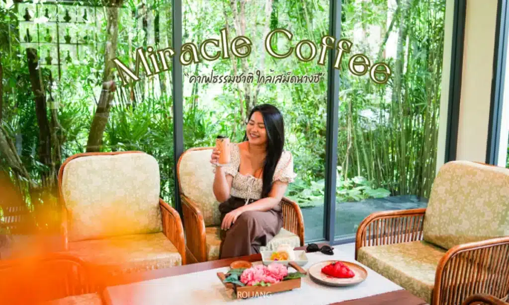 ผู้หญิงคนหนึ่งนั่งบนเก้าอี้หวายที่ร้านกาแฟมิราเคิล ถือเครื่องดื่มและยิ้ม พร้อมทั้งมีขนมและจานผลไม้วางอยู่บนโต๊ะตรงหน้าเธอ.