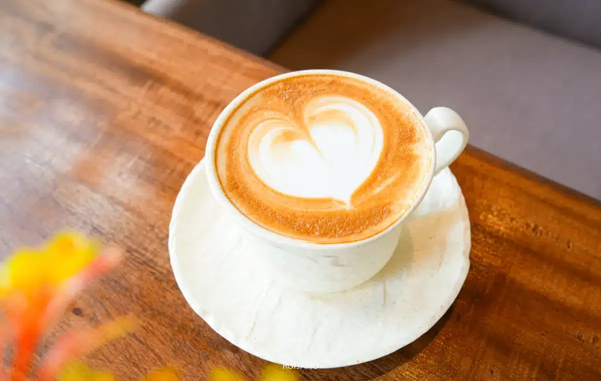 กาแฟคาปูชิโน Miracle Coffee ที่มีลายลาเต้รูปหัวใจวางอยู่บนจานรองสีขาวบนโต๊ะไม้.