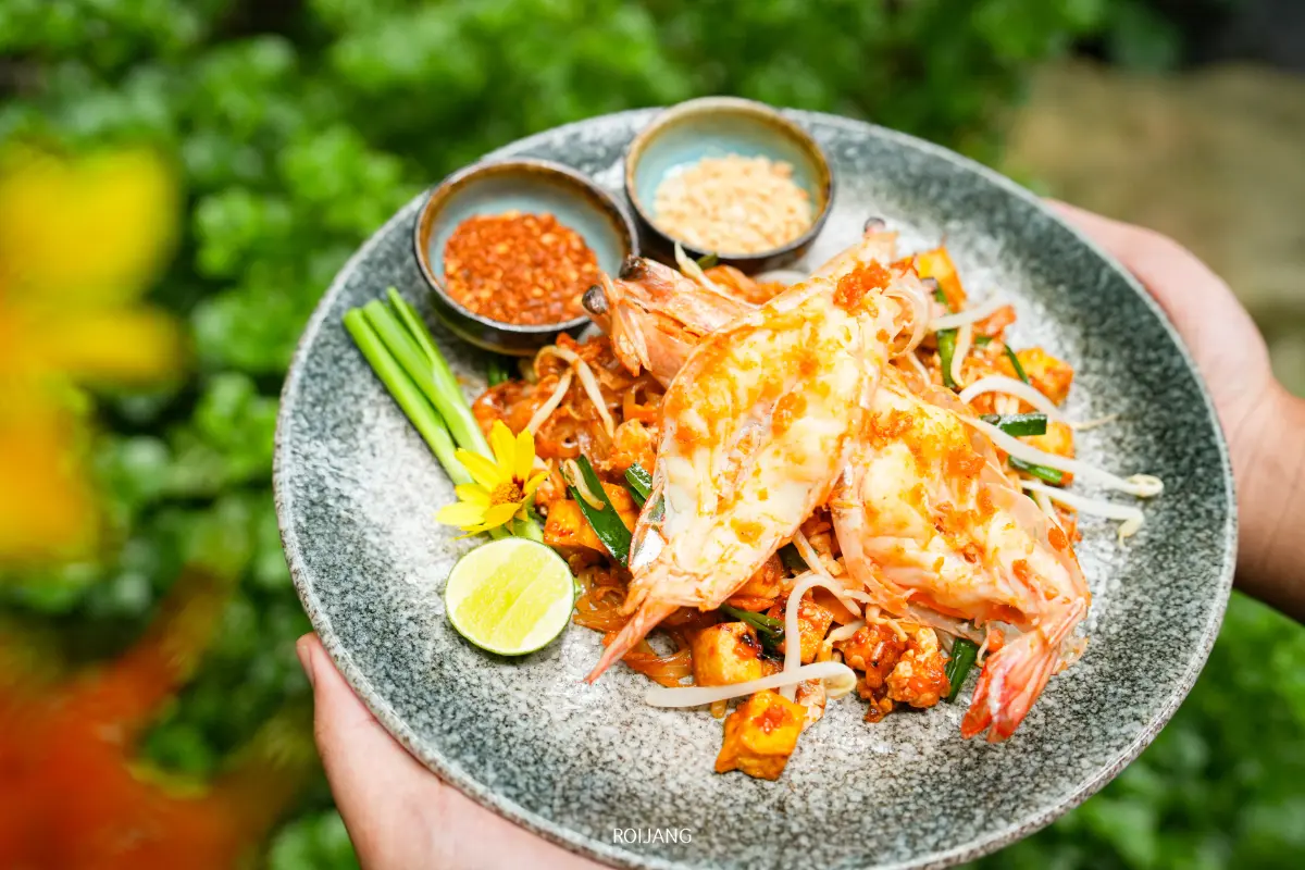 ผัดไทยจานหนึ่งที่มีกุ้งตัวใหญ่สองตัว เสิร์ฟพร้อมมะนาว ต้นหอม ถั่วงอก ถั่วลิสงบด และพริกป่นในชามเล็กๆ ทานคู่กับกาแฟ Miracle Coffee สักแก้ว.