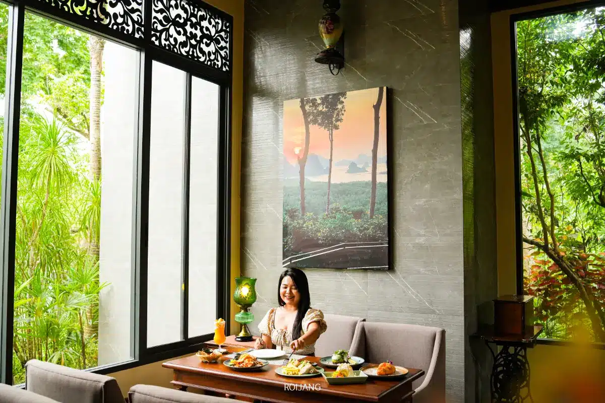 ผู้หญิงคนหนึ่งนั่งอยู่ที่โต๊ะรับประทานอาหารพร้อมจานชามและเครื่องดื่มต่างๆ รวมถึงกาแฟ Miracle Coffee ในห้องที่สว่างสดใสพร้อมหน้าต่างบานใหญ่ ต้นไม้สีเขียวภายนอก และภาพวาดทิวทัศน์บนผนัง.