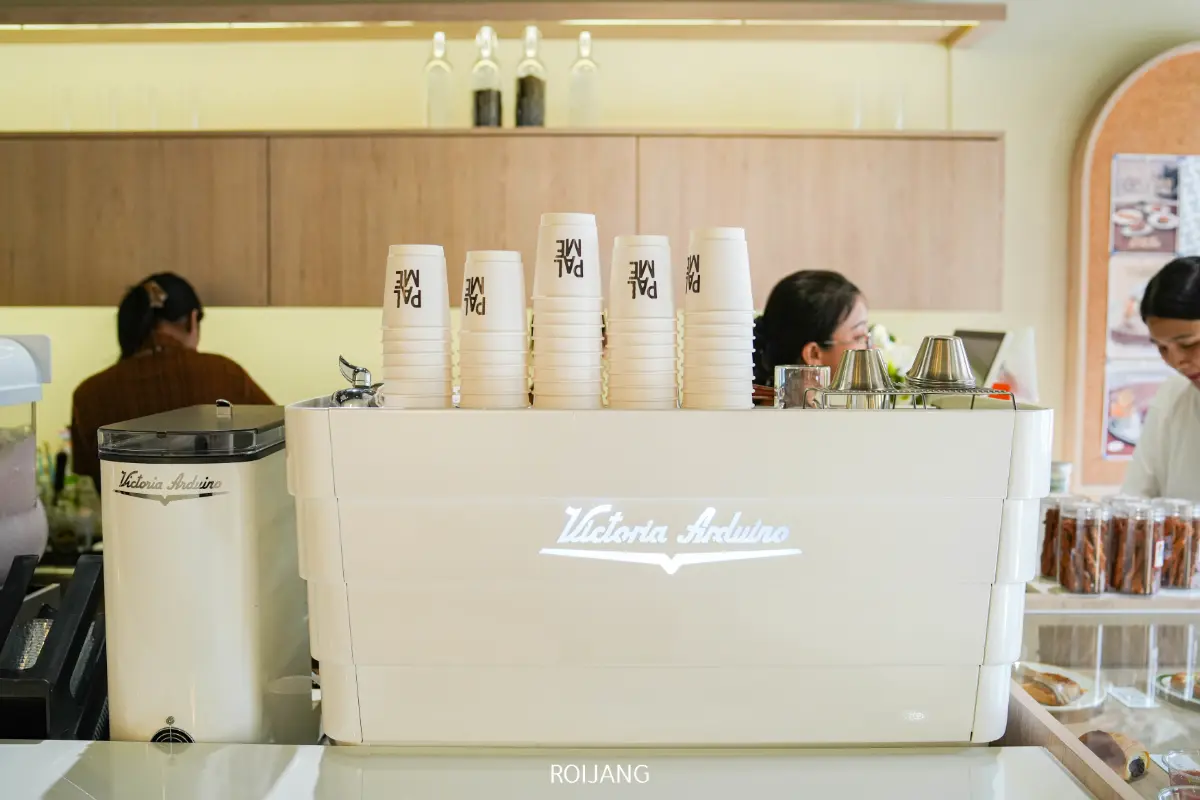 เครื่องชงกาแฟเอสเพรสโซ Victoria Arduino สีขาววางอยู่บนเคาน์เตอร์ข้างแก้วกระดาษ PALME ที่วางซ้อนกัน พนักงาน 2 คนทำงานอยู่หลังเคาน์เตอร์ในบรรยากาศร้านกาแฟที่คึกคัก.