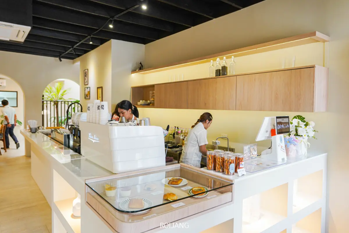 คนสองคนทำงานอยู่ด้านหลังเคาน์เตอร์ร้านกาแฟ PALME ที่ดูทันสมัยและสดใส โดยมีเครื่องชงกาแฟ ขนมอบที่จัดแสดง และอุปกรณ์ต่างๆ ที่จัดวางอย่างเป็นระเบียบ.