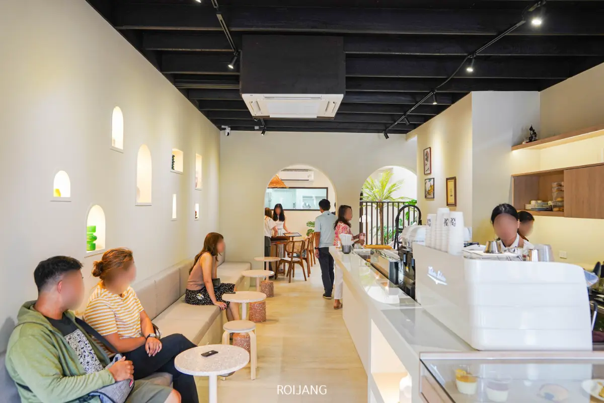 ลูกค้านั่งและยืนอยู่ในร้านกาแฟสไตล์โมเดิร์นมินิมอลที่มีชื่อว่า PALME ซึ่งมีผนังสีอ่อน เคาน์เตอร์สีขาว และบาริสต้าที่กำลังเตรียมเครื่องดื่ม.