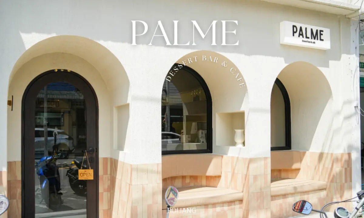 ภายนอกของ PALME Dessert Bar & Café มีหน้าต่างโค้ง ป้ายต้อนรับ "เปิด" ที่ประตู และผนังสีอ่อนพร้อมสีแทนที่ช่วยเน้นให้บรรยากาศอันน่าดึงดูดของ PALME.