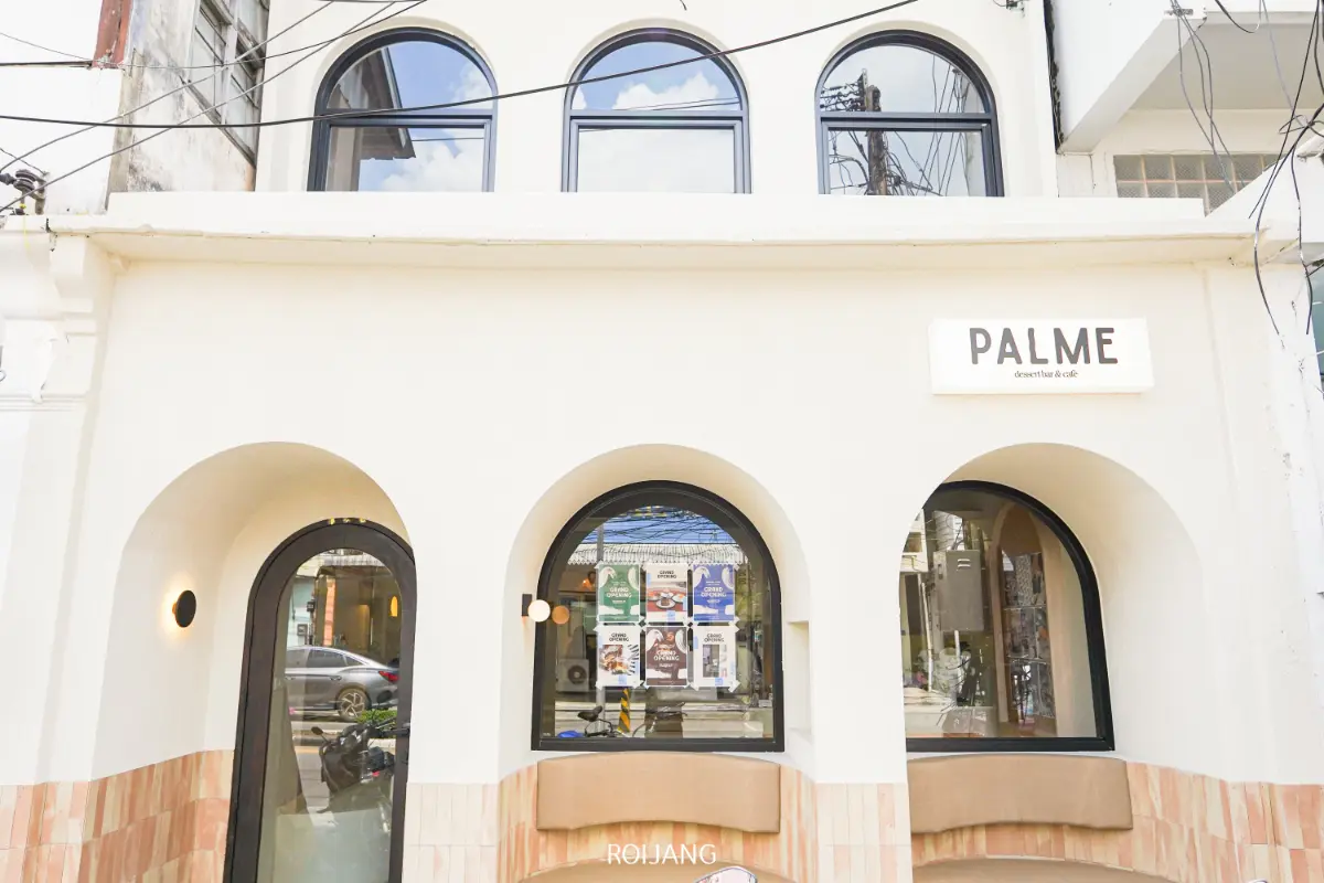 อาคารสีขาวมีหน้าต่างและประตูโค้งสามบาน มีป้ายเขียนว่า "PALME" อยู่เหนือทางเข้า มีโปสเตอร์ PALME ติดไว้ที่หน้าต่างตรงกลาง.