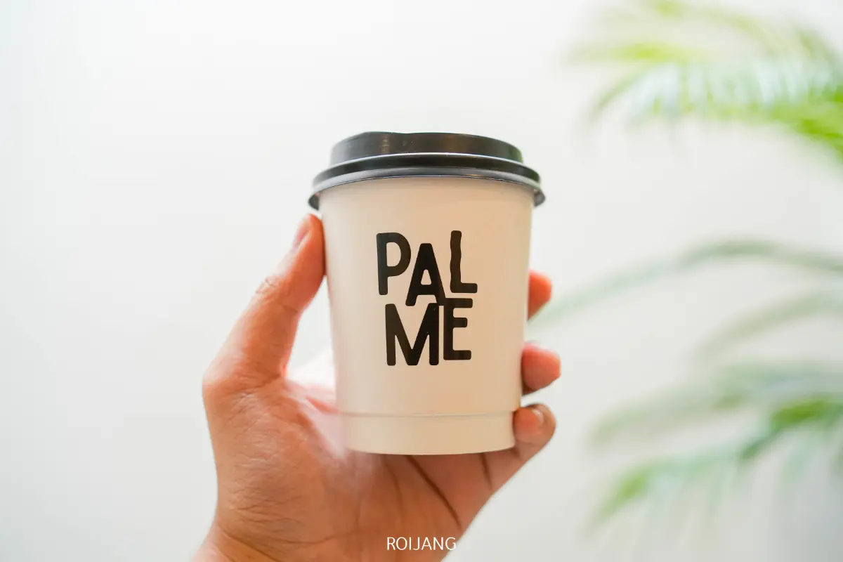 มือข้างหนึ่งถือถ้วยกาแฟกระดาษสีขาวที่มีฝาปิดสีดำ มีตัวอักษรสีดำตัวหนาพิมพ์ว่า “PALME” บนถ้วยกาแฟ ต้นไม้สีเขียวเบลออย่างนุ่มนวลในพื้นหลัง.