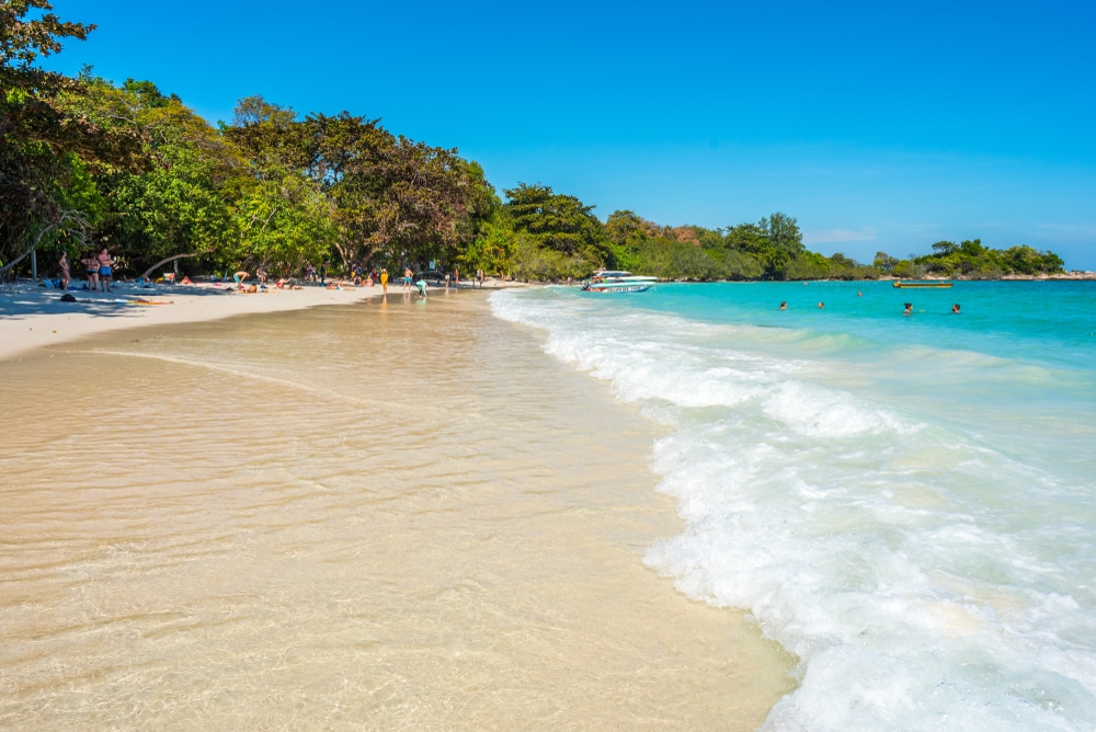 น้ำทะเลสีฟ้าใสและคลื่นทะเลอ่อนๆ ซัดเข้าสู่ชายหาดทรายที่เรียงรายไปด้วยต้นไม้ ผู้คนลงเล่นน้ำและพักผ่อนบนชายหาดภายใต้ท้องฟ้าสดใสและมีแดด เหมาะสำหรับผู้ที่ต้องการ เที่ยวเกาะเสม็ด 