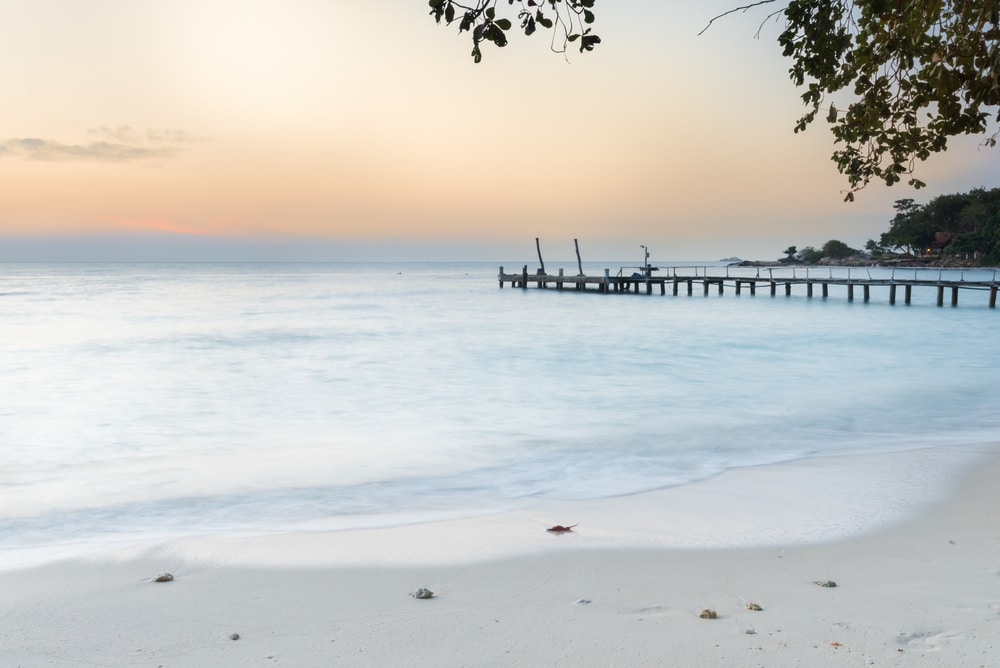 ชายหาดอันเงียบสงบยามพระอาทิตย์ตกดิน พร้อมด้วยคลื่นที่อ่อนโยน หาดทราย เปลือกหอยที่กระจัดกระจาย  ที่เที่ยวเกาะเสม็ด และท่าเรือไม้ที่ยื่นออกไปเหนือผืนน้ำ เหมาะสำหรับผู้ที่เที่ยวเกาะเสม็ด.