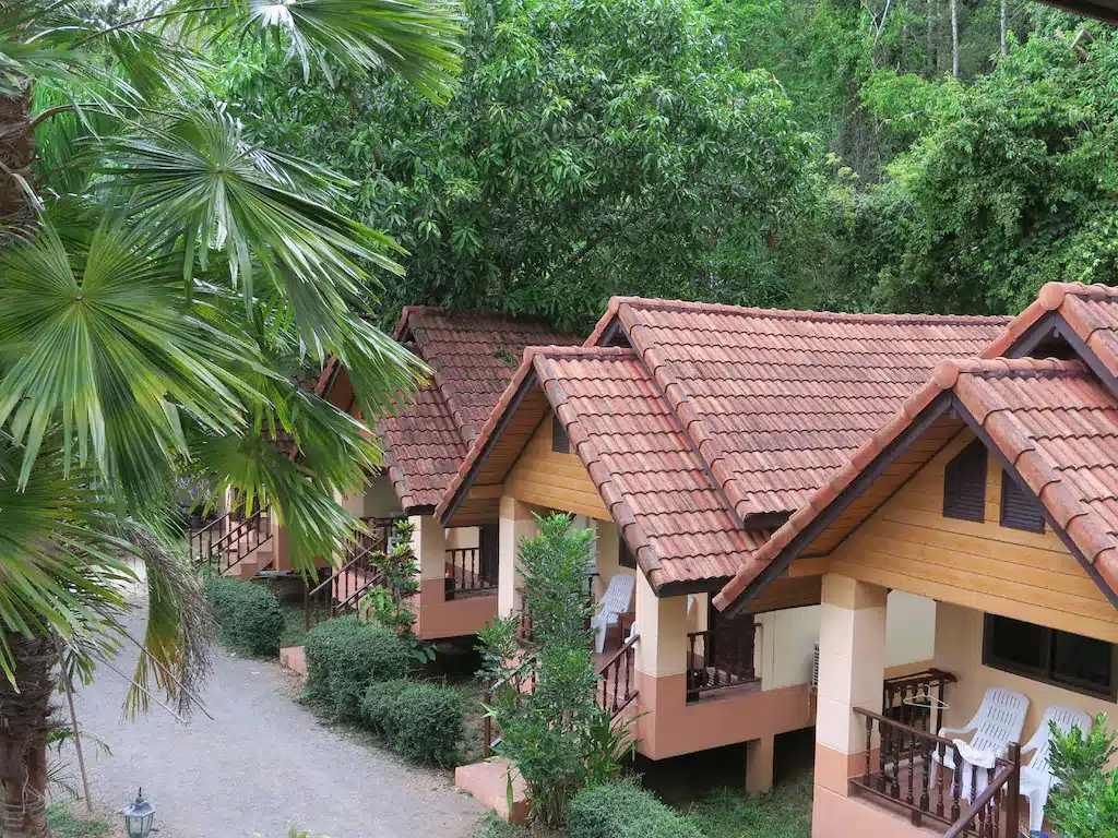 กระท่อมเล็กๆ เรียงเป็นแถวหลังคากระเบื้องสีแดง มีระเบียงและเก้าอี้  ที่พักสตูลแนวธรรมชาติ ล้อมรอบด้วยต้นไม้เขียวขจีและทางเดินลูกรัง เหมาะสำหรับผู้ที่แสวงหาสตูลแนวธรรมชาติ.
