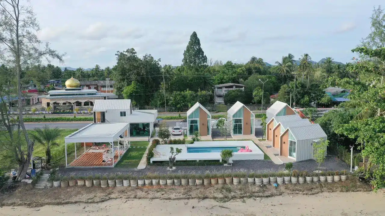 มุมมองทางอากาศของที่พักริมชายหาดที่ทันสมัย พร้อมด้วยบ้านหลังเล็ก สระว่ายน้ำ ลานบ้าน สนามหญ้าสี ที่พักสตูลแนวธรรมชาติ เขียว และกลิ่นอายสตูลแนวธรรมชาติ ตั้งอยู่ใกล้ถนนที่มีต้นไม้และอาคารเป็นฉากหลัง.