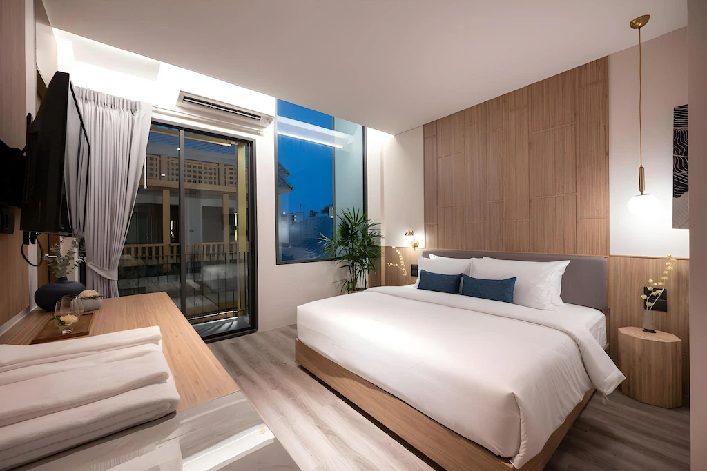 ห้องพักในโรงแรมทันสมัยพร้อมเตียงขนาดใหญ่ เฟอร์นิเจอร์ไม้ ทีวีติดผนัง โต๊ะทำงาน และหน้าต่างบานใหญ่ที่มองเห็นระเบียงในยามพลบค่ำ.