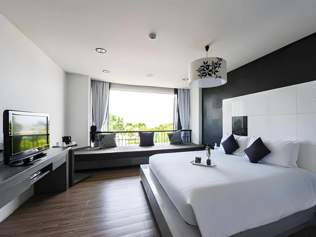 ห้องพักโรงแรมสมัยใหม่พร้อมเตียงขนาดใหญ่ การตกแต่งสีดำและขาว ทีวีจอแบน ที่นั่งริมหน้าต่างยาว โต๊ะทำงาน และหน้าต่างพร้อมผ้าม่านที่มองเห็นวิวสีเขียว—เหมาะสำหรับผู้ที่ต้องการที่พักชะอำที่สะดวกสบายและมีสไตล์.