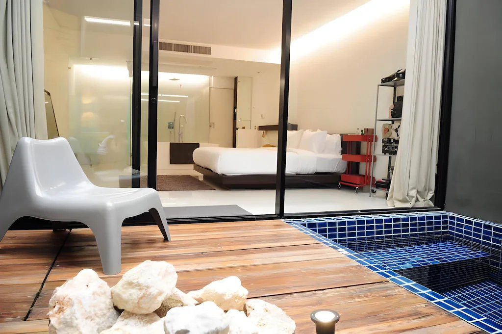 ห้องพักในโรงแรมทันสมัยพร้อมเตียงคู่ มองผ่านประตูบานเลื่อนกระจกจากลานที่มีสระว่ายน้ำเล็กที่ปูด้วยกระเบื้อง เก้าอี้พลาสติก และหินบนพื้นไม้.