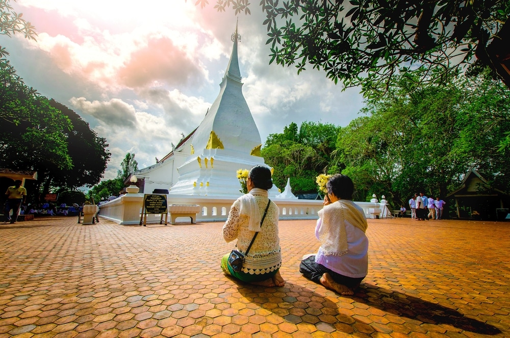 สองคนคุกเข่าและภาวนาอยู่หน้าพระธาตุสีขาวในลานปูพื้นด้วยหิน ต้นไม้ล้อมรอบ ท้องฟ้าครึ่งหนึ่งมีเมฆ ที่เที่ยวเลย ปกคลุม—ภาพที่น่าใจชื่นสำหรับใครก็ตามที่กำลังสำรวจ เที่ยวเลย.