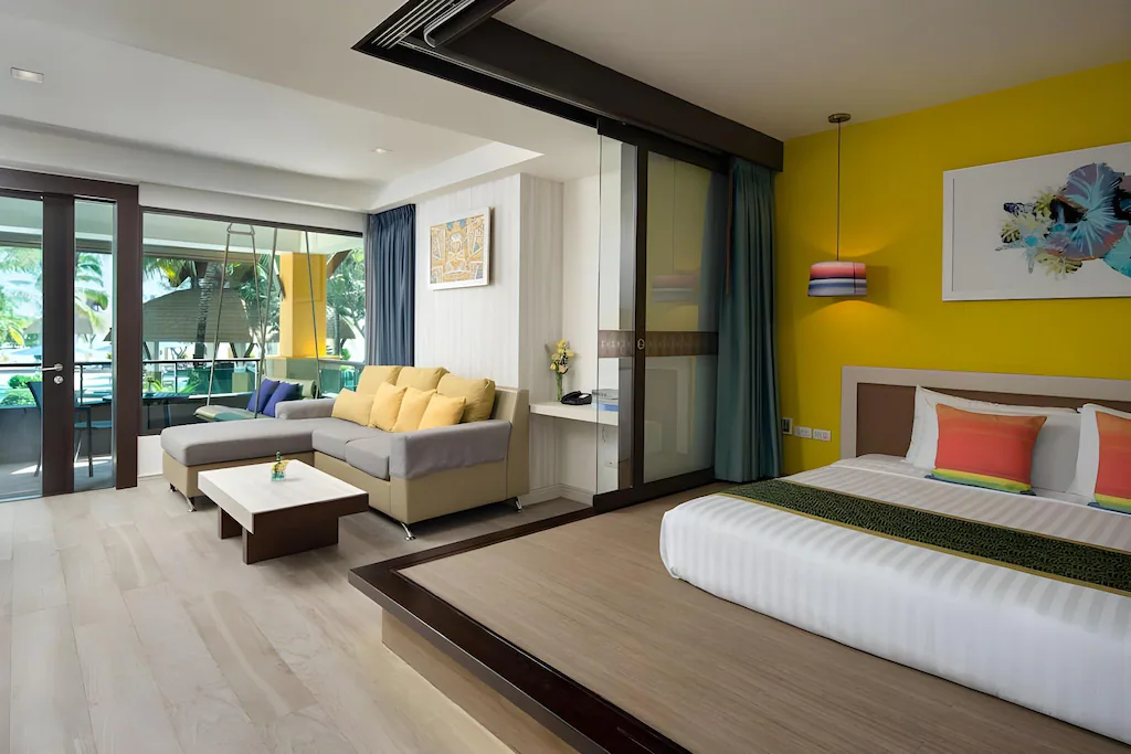 ห้องพักโรงแรมสมัยใหม่พร้อมเตียงคู่, รายละเอียดสีสันสดใส, โซฟา, โต๊ะกาแฟ, และระเบียงพร้อมที่นั่งกลางแจ้งที่สามารถมองเห็นต้นปาล์ม—เป็นตัวเลือกที่สมบูรณ์แบบสำหรับผู้ที่ต้องการ โรงแรมชะอำ หรือ ที่พักชะอำ เพื่อเพลิดเพลินกับความสะดวกสบายและวิวทิวทัศน์ที่สวยงาม.