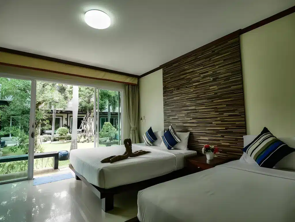 ห้องพักโรงแรมพร้อมเตียงสองเตียง ผนังตกแต่งลายทาง ประตูบานเลื่อนกระจก และผ้าขนหนูพับเป็นรูปหงส์—เหมาะสำหรับการพักผ่อนและเพลิดเพลินกับวิวสวนด้านนอก เหมาะสำหรับผู้ที่มองหาที่พักชะอำหรือที่พักชะอำราคาประหยัด.