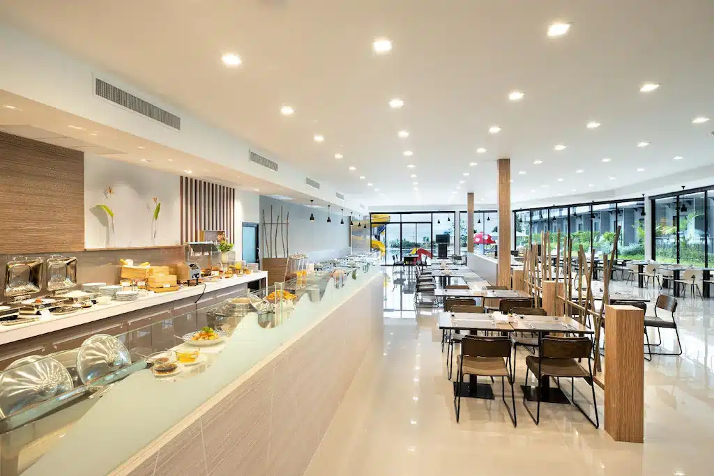 ร้านอาหารบุฟเฟ่ต์ที่สว่างและทันสมัย มีเคาน์เตอร์อาหารยาวอยู่ทางซ้าย มีจานอาหารที่ปิดฝาไว้เรียบร้อย และโต๊ะอาหารที่จัดเรียงอย่างเป็นระเบียบบนพื้นเงางาม—เหมาะสำหรับแขกที่เข้าพักที่ โรงแรมชะอำ หน้าต่างบานใหญ่เรียงรายอยู่ทางด้านขวา.