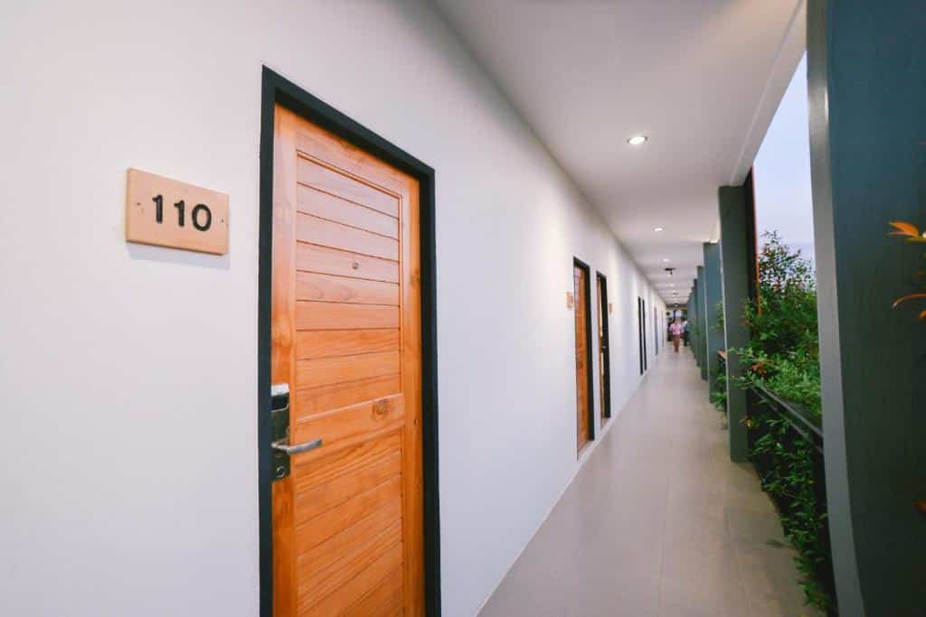 ทางเดินที่มีประตูไม้หลายบาน รวมถึงบานหนึ่งที่มีป้ายเขียนว่า "110" ผนังสีขาว และต้นไม้เรียงรายอยู่ทางด้าน เที่ยวเลย ขวา—ให้ความรู้สึกอบอุ่นเหมือนบรรยากาศของ ที่เที่ยวเลย.