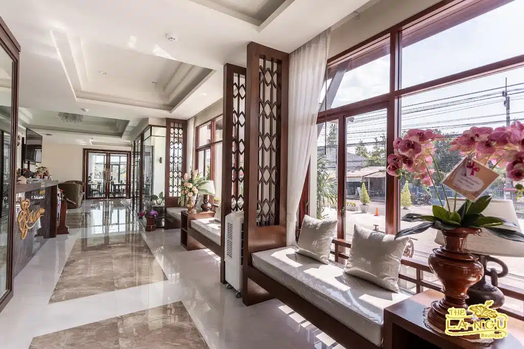 ล็อบบี้ของโรงแรมสว่างสดใสพร้อม ที่พักสตูลแนวธรรมชาติ หน้าต่างบานใหญ่ ตกแต่งด้วยไม้ ม้านั่งบุนวม กล้วยไม้ในกระถาง และพื้นหินอ่อน แสงแดดส่องเข้ามาสร้างบรรยากาศอบอุ่นชวนให้นึกถึงการใช้เวลาสตูลแนวธรรมชาติ.