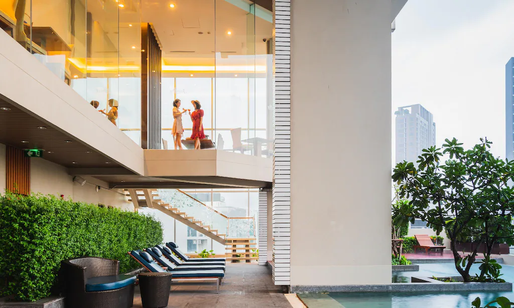 อาคารทันสมัยที่มีผนังกระจก ผู้คนเดินอยู่ข้างในที่ชั้นบน เก้าอี้เลานจ์ริมสระน้ำ และต้นไม้สีเขียวตามชั้นล่าง.