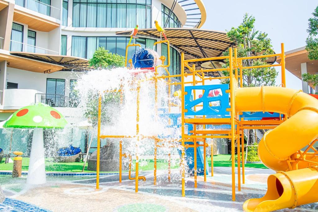 สนามเด็กเล่นน้ำกลางแจ้งพร้อมสไลด์สีเหลือง, ฟีเจอร์น้ำกระเซ็น, และโครงสร้างปีนป่ายอยู่หน้าตึกสไตล์โมเดิร์น—เหมาะสำหรับครอบครัวที่มองหาความสนุกใกล้ที่พักชะอำหรือโรงแรมชะอำในวันที่แดดออก.