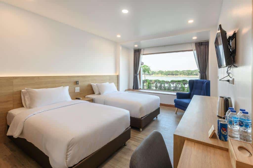 ห้องพักโรงแรมสมัยใหม่พร้อมเตียง ที่เที่ยวเลย เดี่ยวสองเตียง หน้าต่างบานใหญ่พร้อมวิว—เหมาะสำหรับการพักผ่อนหลังจากเที่ยวมา—โต๊ะทำงานพร้อมน้ำดื่มบรรจุขวด ทีวีติดผนัง และเก้าอี้มีแขนสีน้ำเงินตั้งอยู่ข้างหน้าต่าง.