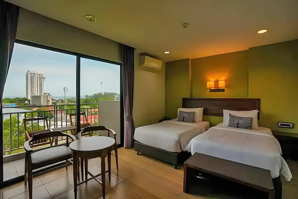 ห้องพักในโรงแรมที่มีเตียงคู่ 2 เตียง โต๊ะและเก้าอี้ตัวเล็ก พื้นไม้ หน้าต่างบานใหญ่ และระเบียงที่มองเห็นวิวเมือง เหมาะสำหรับนักเดินทางที่กำลังมองหาที่พักผ่อนสัตหีบหลังจากเที่ยวที่เที่ยวสัตหีบแล้ว.