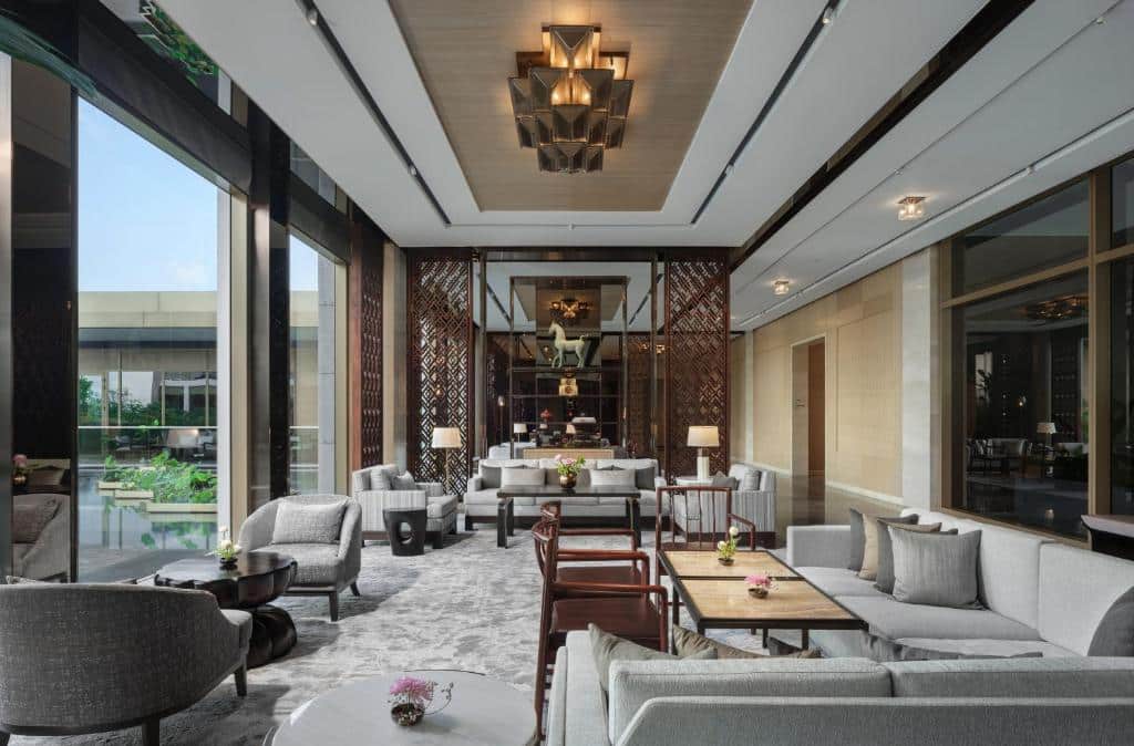 เลานจ์โรงแรมทันสมัยพร้อมหน้าต่างบานใหญ่ โซฟาและเก้าอี้โทนสีกลางๆ โต๊ะไม้ ไฟประดับ และต้นไม้ในร่ม แสงธรรมชาติส่องเข้ามาเติมเต็มพื้นที่.
