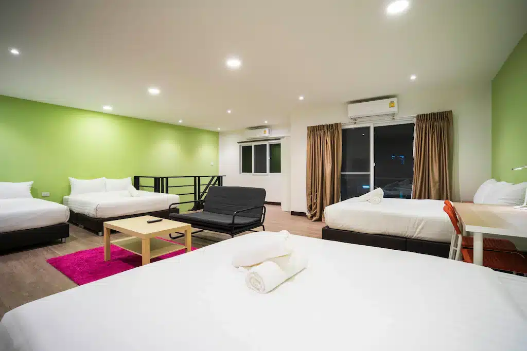 ห้องพักในโรงแรมที่สดใสและทันสมัยมีเตียงสี่เตียง โซฟาสีดำ โต๊ะกาแฟ โต๊ะทำงานพร้อมเก้าอี้สีส้ม หน้าต่างบานใหญ่ ผนังสีเขียว และพื้นไม้.
