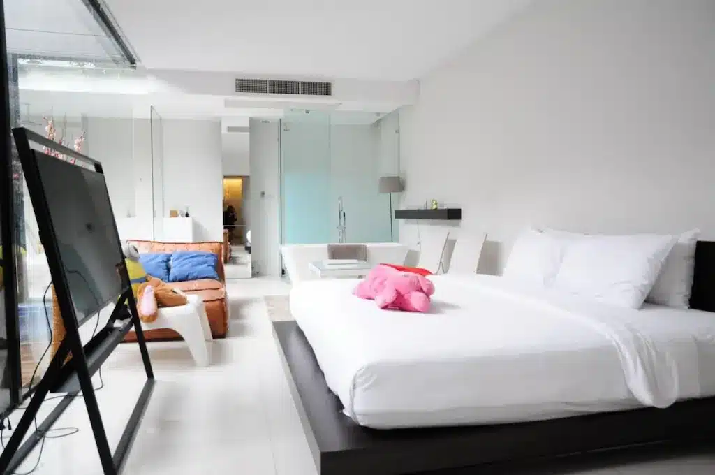 ห้องนอนทันสมัยพร้อมเตียงขนาดใหญ่ ชุดเครื่องนอนสีขาว ตุ๊กตาสัตว์สีชมพู ทีวีจอแบน ฉากกั้นกระจก และพื้นที่นั่งเล่นพร้อมโซฟาสีน้ำตาล.