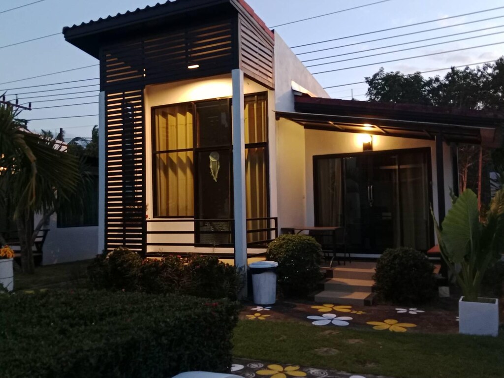 บ้านหลังเล็กสไตล์โมเดิร์นที่มีหน้าต่างบานใหญ่ ระเบียงหน้าบ้าน สวนที่ ที่พักสตูลแนวธรรมชาติ มีบันไดหินรูปดอกไม้ และไฟภายนอกอาคารในยามพลบค่ำ เหมาะสำหรับผู้ที่กำลังมองหาที่พักสไตล์ธรรมชาติ.