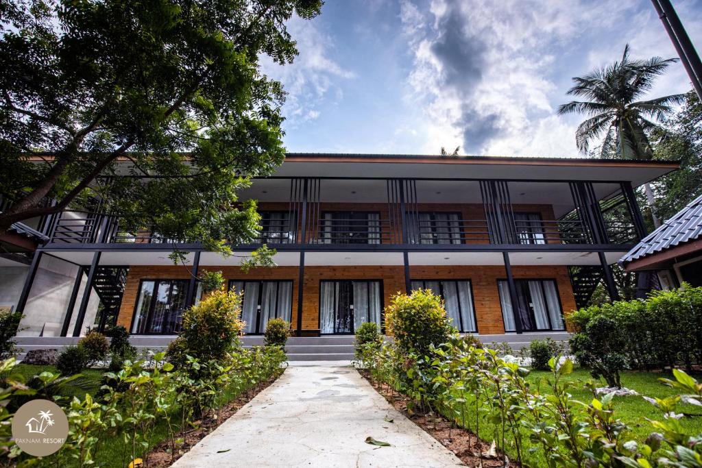 อาคารรีสอร์ททันสมัย 2 ชั้นพร้อม ที่พักสตูลแนวธรรมชาติ ระเบียง หน้าต่างบานใหญ่ และทางเดินที่รายล้อมไปด้วยแมกไม้เขียวขจีและพืชพรรณนานาชนิดภายใต้ท้องฟ้าที่มีเมฆเป็นบางส่วน เหมาะสำหรับผู้ที่กำลังมองหาสตูลแนวธรรมชาติ.