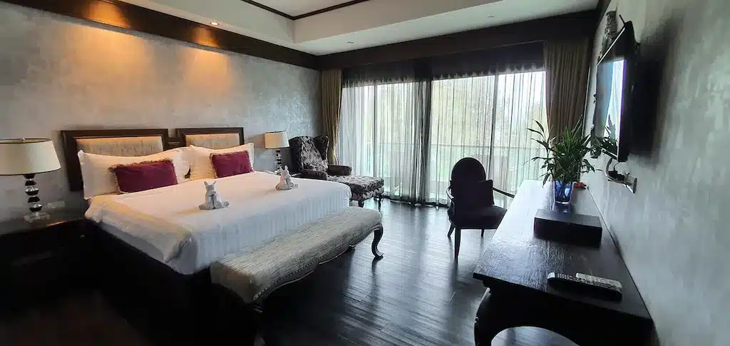 ห้องพักโรงแรมกว้างขวางที่โรงแรมชะอำ พร้อมเตียงขนาดใหญ่ โคมไฟ ม้านั่ง เก้าอี้ โต๊ะทำงานพร้อมต้นไม้ ทีวีติดผนัง และหน้าต่างบานใหญ่จากพื้นจรดเพดานพร้อมผ้าม่าน มองเห็นวิวสีเขียวขจี เหมาะสำหรับผู้ที่มองหาที่พักชะอำราคาประหยัด.