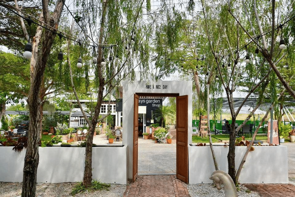 ประตูไม้แบบเปิดนำไปสู่ร้านค้าในสวนที่มีเสน่ห์ในที่เที่ยวสัตหีบ ซึ่งเต็มไปด้วยไม้กระถาง ต้นไม้ และป้ายเขียนว่า "ขอให้มีความสุขนะ" เหนือทางเข้า.