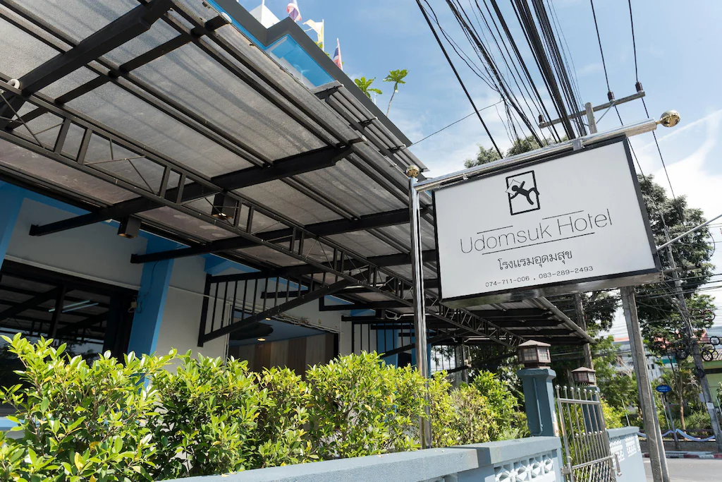 วิวภายนอกโรงแรมอุดมสุข ที่พักสไตล์ ที่พักสตูลแนวธรรมชาติ ธรรมชาติ มีป้ายสีขาว เขียวขจีริมรั้ว และส่วนหนึ่งของตัวอาคารและกันสาดใต้ฟ้าใส.