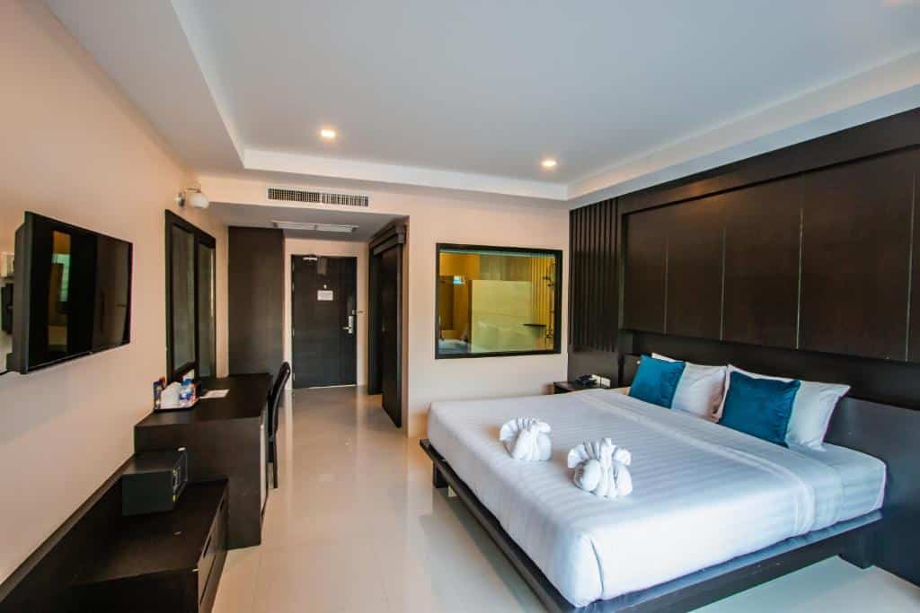 ห้องพักโรงแรมสมัยใหม่พร้อมเตียงขนาดใหญ่ ผ้าปูที่นอนสีขาว หมอนอิงสีน้ำเงิน แต่งด้วยศิลปะผ้าขนหนู โต๊ะทำงาน ทีวีติดผนัง และหน้าต่างที่มองเห็นห้องน้ำ—เหมาะสำหรับนักเดินทางที่กำลังมองหาที่พักชะอำราคาประหยัดในสไตล์ที่ทันสมัย.