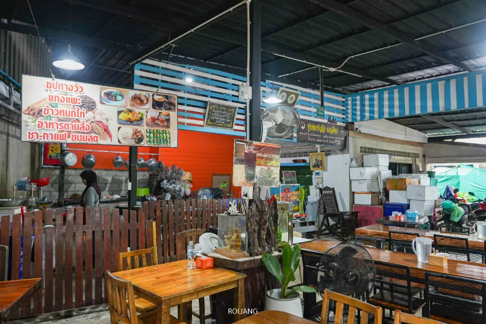 ภายในร้านอาหารไทยบรรยากาศสบายๆ มีโต๊ะและเก้าอี้ไม้ เคาน์เตอร์พร้อมอุปกรณ์ทำครัว เมนูบนผนังที่มีรายการพิเศษ เช่น ซุปหางวัวมาลิตา และของตกแต่งลายทางสีฟ้าและขาว.