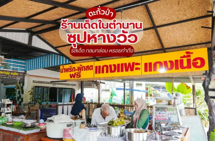 แผงขายอาหารท้องถิ่นที่มีป้ายสีเหลืองจำหน่ายน้ำพริกเผา ผัก แกง เมนูเนื้อ และซุปหางวัวมาลิตาพิเศษฟรี มีคน 2 คนกำลังเตรียมอาหารอยู่หลังเคาน์เตอร์.