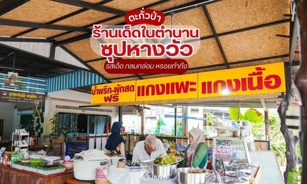 แผงขายอาหารท้องถิ่นที่มีป้ายสีเหลืองจำหน่ายน้ำพริกเผา ผัก แกง เมนูเนื้อ และซุปหางวัวมาลิตาพิเศษฟรี มีคน 2 คนกำลังเตรียมอาหารอยู่หลังเคาน์เตอร์.