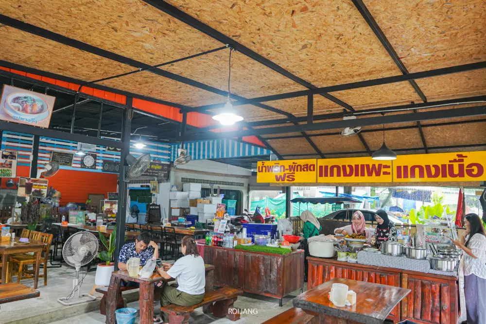ร้านอาหารไทยบรรยากาศสบายๆ มีโต๊ะไม้ คนนั่งทานอาหาร และมีเคาน์เตอร์ใส่หม้อใหญ่ๆ ใส่ซุปหางวัวมาลิตา มีป้ายสีเหลืองเขียนภาษาไทยให้เห็นเหนือเคาน์เตอร์.