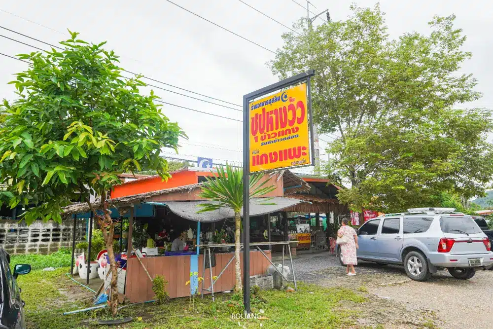 ร้านอาหารริมถนนหลังคาสีส้ม ที่นั่งกลางแจ้ง ป้ายไทยสีเหลืองโฆษณาหางวัวมาลิตา รถจอด ต้นไม้ และท้องฟ้ามีเมฆมาก.