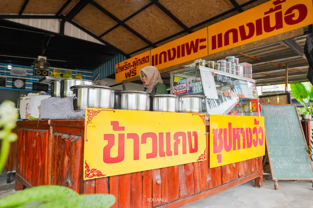ผู้หญิงคนหนึ่งยืนอยู่ด้านหลังแผงขายอาหารที่ทำจากไม้พร้อมกระถางขนาดใหญ่และป้ายไทยสีเหลืองสดใสที่โฆษณาหางวัวมาลิตาใต้โครงสร้างหลังคา.