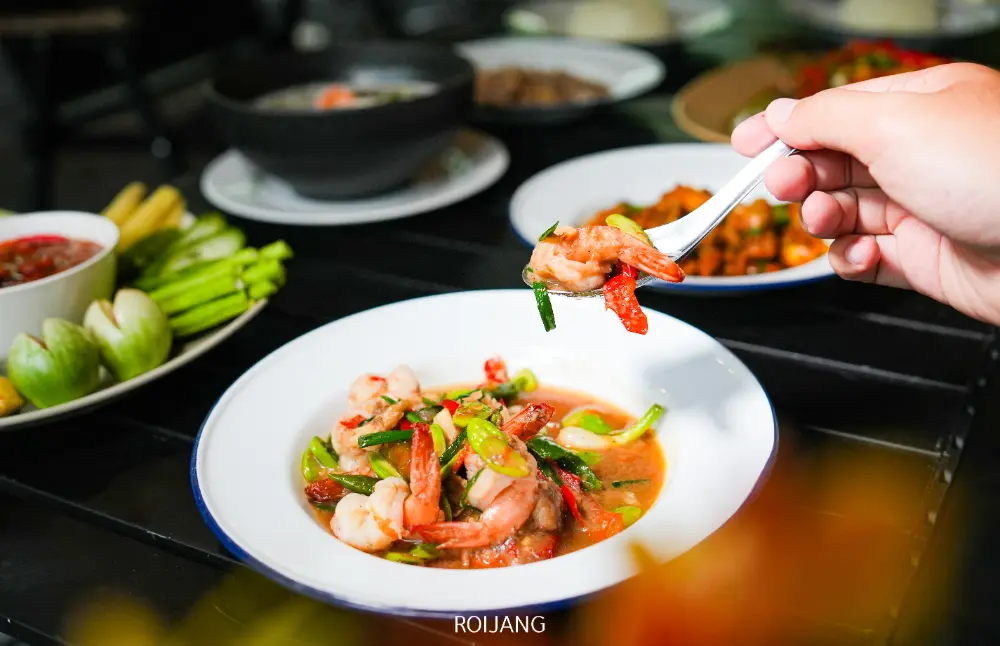 มือหนึ่งถือส้อมที่มีกุ้งและผักอยู่เหนือจานผัดกุ้งสีขาว ล้อมรอบด้วยอาหารอื่นๆ บนโต๊ะมืดที่ท้ายสุขภาพดีแคมป์ปิ้ง.