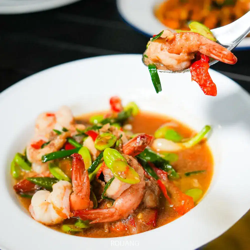 ชามสีขาวไส้กุ้ง ผักใบเขียว และพริกแดงในซอสสีน้ำตาลที่ท้ายแคมป์ปิ้ง ช้อนยกส่วนหนึ่งของจานที่มีรสชาติไว้เหนือชาม.