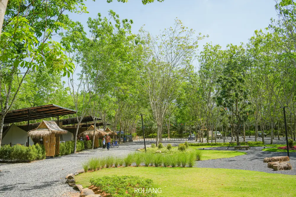 พื้นที่กลางแจ้งที่มีภูมิทัศน์สวยงามที่ท้ายแคมป์ปิ้งมีสนามหญ้าสีเขียว ทางเดินลูกรัง ต้นไม้กระจัดกระจาย กระท่อมหลังเล็ก และคนสองสามคนกำลังเดินอยู่ใต้ท้องฟ้าแจ่มใส.