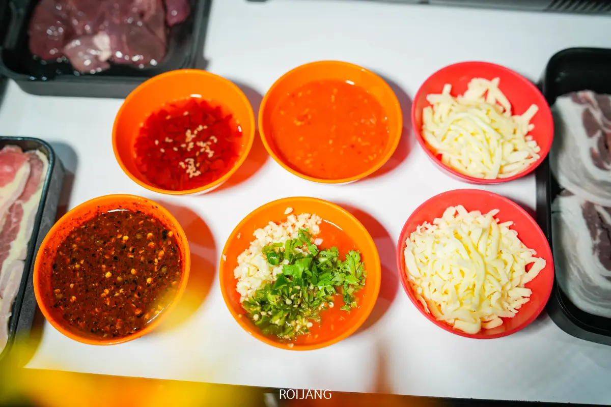 ชามสีส้มหกใบที่บรรจุซอส, สมุนไพรสับ, และชีสขูดฝอยถูกจัดเรียงบนโต๊ะข้างถาดเนื้อดิบที่ สถานีดีจัง อันคึกคัก.